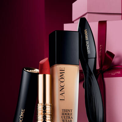 Un elegante bodegon con la barra de labios mate L'Absolu Rouge de Lancome, la base de maquillaje Teint Idole Ultra Wear y la mascara Hypnose Drama sobre un lujoso fondo navideno en tonos rojo oscuro.