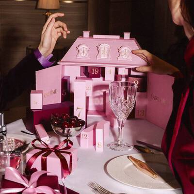 Manos interactuando con un calendario de adviento rosa Lancome sobre una mesa festiva con vajilla y cerezas.