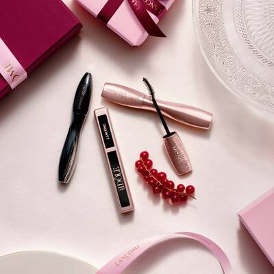 Un arreglo flat-lay de productos de maquillaje Lancome, con mascara de pestanas y brillo de labios, junto a cajas de regalo rosas, lazos de saten y una ramita de grosellas rojas.
