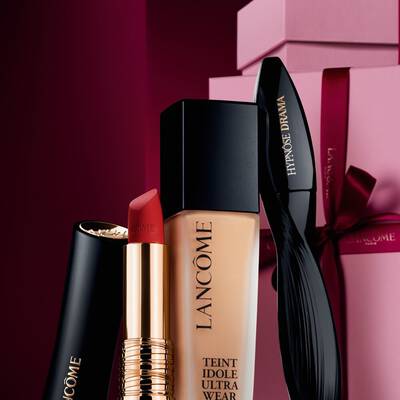 Una elegante naturaleza muerta de pintalabios mate L'Absolu Rouge de Lancome, base de maquillaje Teint Idole Ultra Wear y mascara Hypnose Drama sobre un lujoso fondo navideno rojo oscuro.