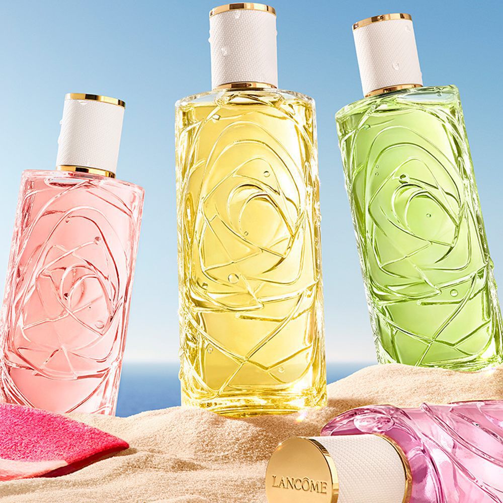 Escena de playa con tres botellas de perfume verticales (rosa, amarillo, verde) y una botella rosa tirada en la arena. A la izquierda hay una toalla con rayas rosas con un cielo azul claro y un horizonte oceanico de fondo.
