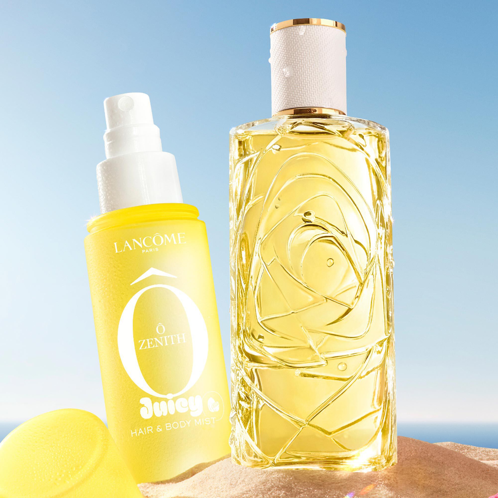 Dos botellas amarillas, una botella rectangular de perfume con un diseno texturizado y otra botella de spray con un tapon amarillo en el lateral, situada en una playa de arena. Una toalla de playa doblada de rayas rosas esta junto a las botellas. Al fondo, hay un cielo azul despejado y el oceano.