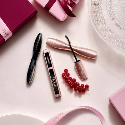 Una composicion flat-lay de productos de maquillaje Lancome —con mascara de pestanas y brillo de labios— acompanada de cofres de regalo rosas, lazos de saten y una ramita de bayas rojas.