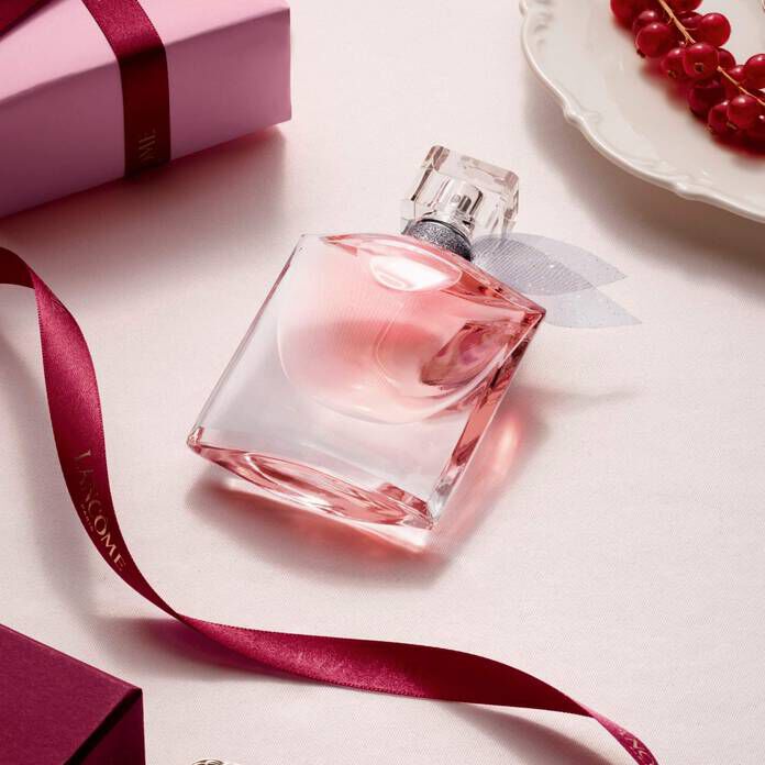 Un frasco rosa de perfume Lancome decorado sobre una superficie clara con una caja de regalo, una cinta y grosellas rojas.