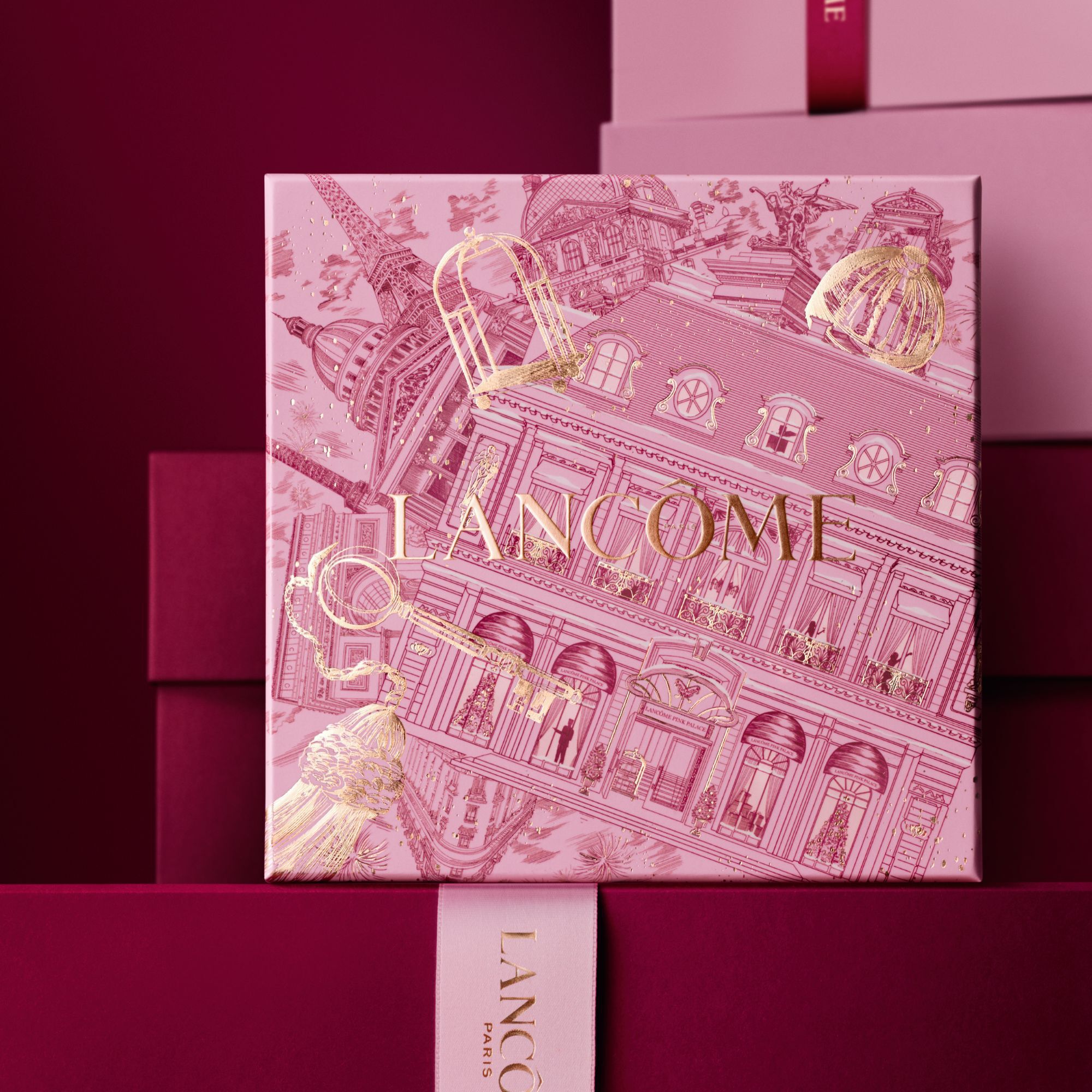 Imagen de una caja de regalo rosa Lancome de pie delante de un monton de cajas de regalo rosas y rojo oscuro, sobre un fondo rojo oscuro.