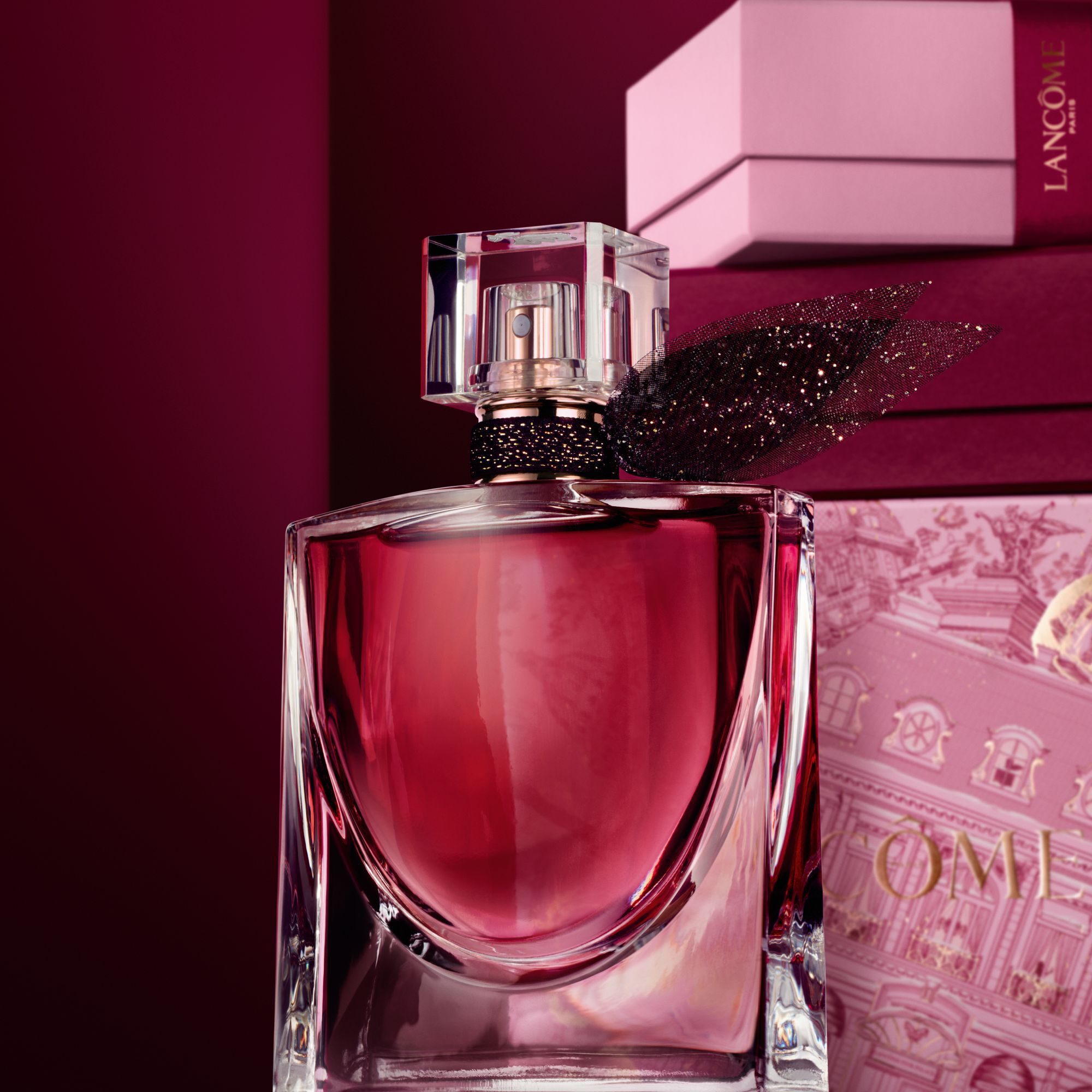 Imagen de La vie est belle L'Elixir de pie frente a un monton de cajas de regalo rosas y rojo oscuro, sobre un fondo rojo oscuro.