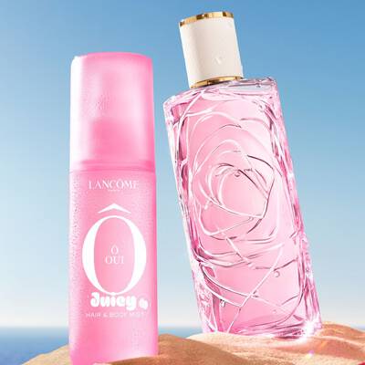 Una escena de playa con tres coloridos frascos de perfume erguidos en la arena, uno rosa, uno amarillo y otro verde, con otro frasco rosa de perfume de lado en primer plano. Una toalla a rayas rosas y blancas esta colocada sobre la arena a la izquierda. El fondo muestra un cielo azul claro que se transforma en un oceano azul en el horizonte.