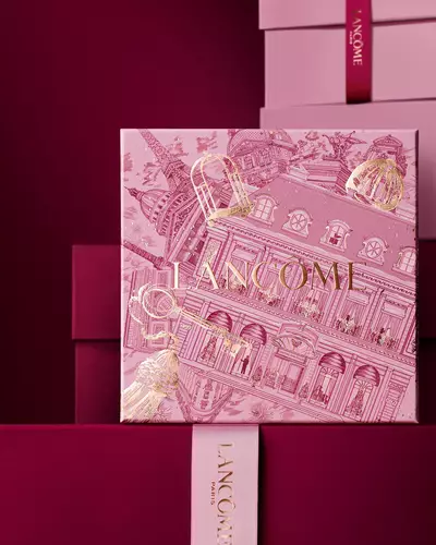 Imagen de una caja de regalo rosa Lancome de pie delante de un monton de cajas de regalo rosas y rojo oscuro, sobre un fondo rojo oscuro.