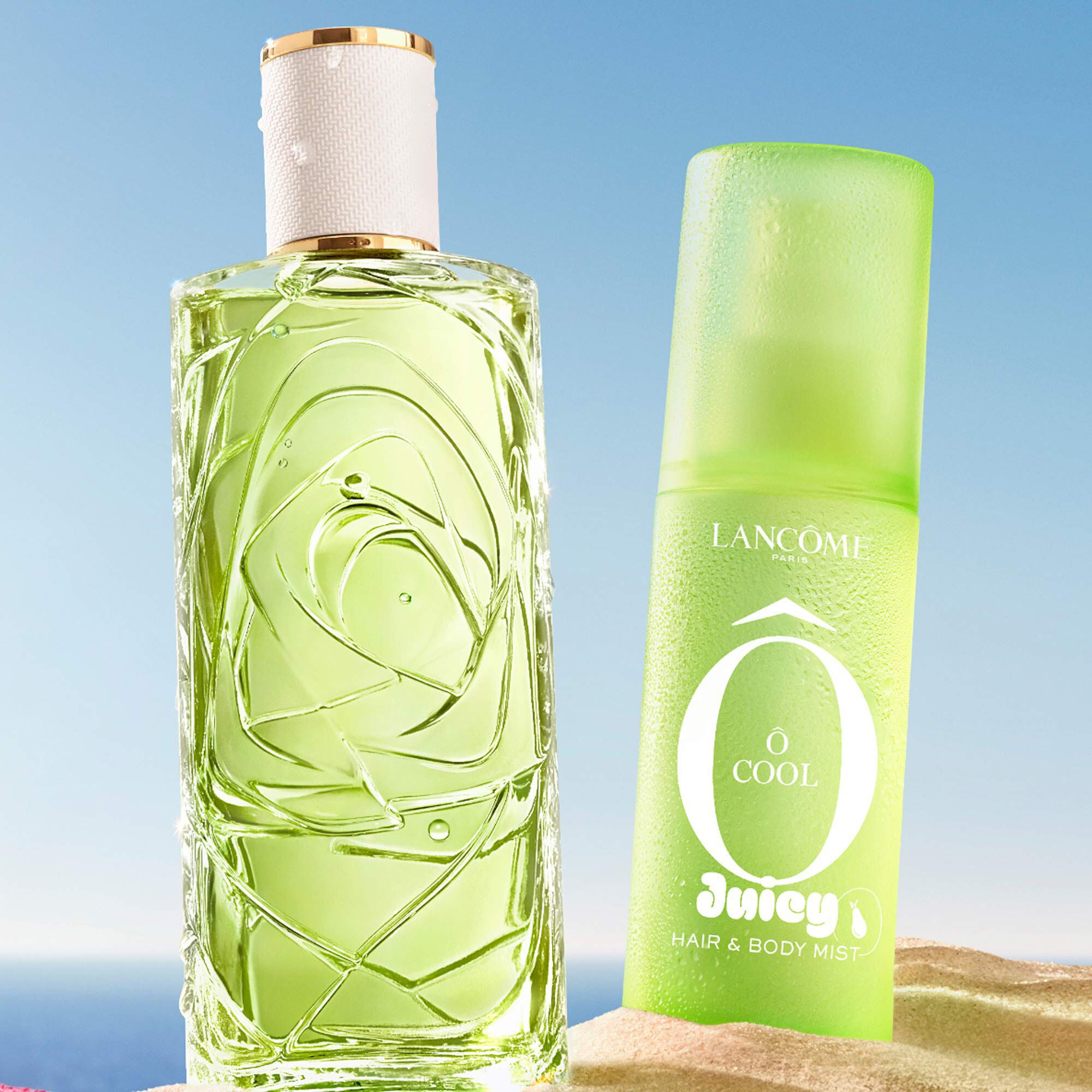 Dos botellas verdes, una botella rectangular de perfume con un diseno texturizado y una botella de spray, situada en una playa arenosa. Una toalla de playa doblada de rayas rosas esta junto a las botellas. Al fondo, hay un cielo azul despejado y el oceano.