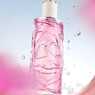 Un frasco de perfume rosa claro con un diseno de vidrio texturizado, sobre un fondo azul claro del cielo, con gotas de agua salpicando alrededor. La botella se ve a traves de una abertura circular en una pared rosa texturizada, y hay flores rosas borrosas en primer plano y fondo.