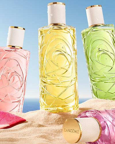 Dos botellas rosas, una botella de cristal transparente con perfume con un diseno texturizado y una botella de spray, situada en una playa arenosa. Una toalla de playa doblada de rayas rosas esta junto a las botellas. Al fondo, hay un cielo azul despejado y el oceano.