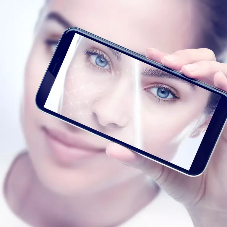 Una persona sosteniendo un telefono movil que muestra un primer plano de su rostro, con enfoque en los ojos y un efecto grafico sobre la piel.