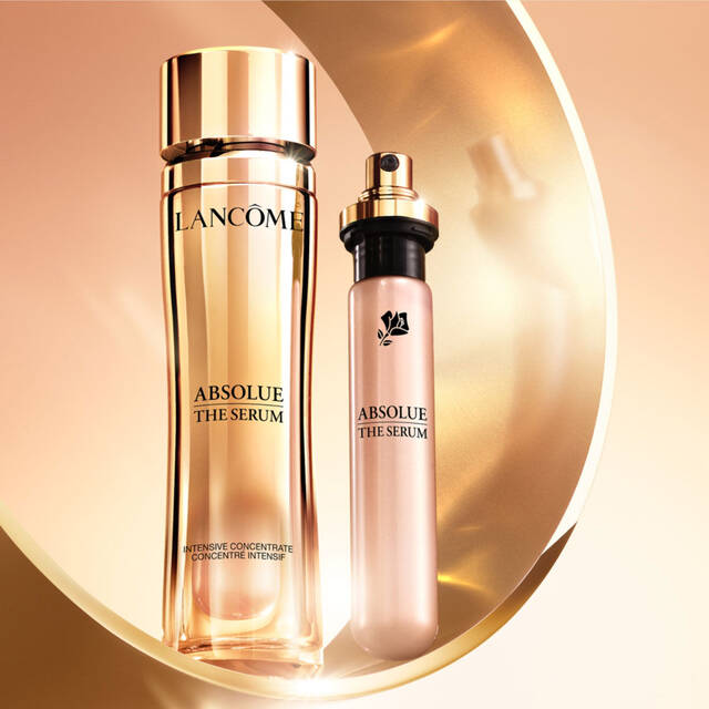 Imagen de dos frascos de serum de la marca Lancome, presentados sobre un fondo dorado. El producto principal es un frasco alto y elegante de "Absolue The Serum", junto a un frasco mas pequeno del mismo producto.