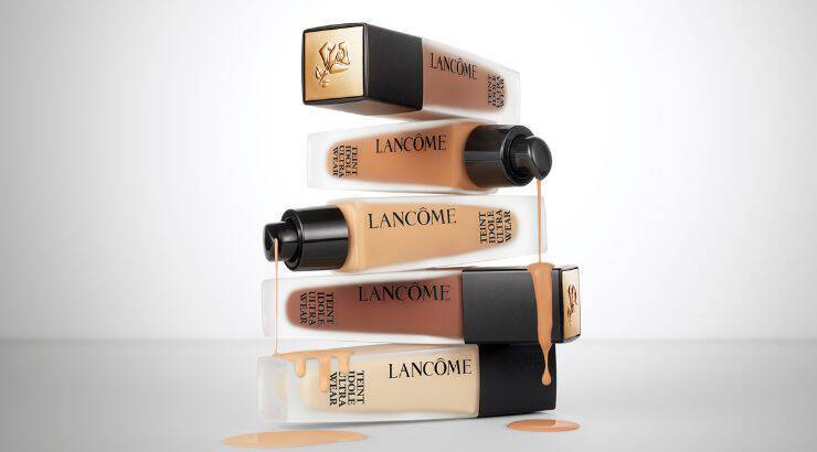 Texto alternativo: Una pila de frascos de base de maquillaje de Lancome, con liquido derramandose de algunos de ellos, sobre un fondo blanco.