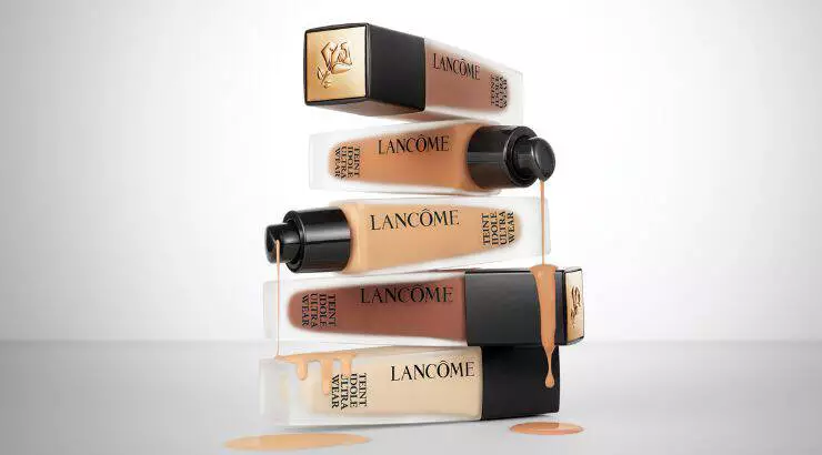 Texto alternativo: Una pila de frascos de base de maquillaje de Lancome, con liquido derramandose de algunos de ellos, sobre un fondo blanco.