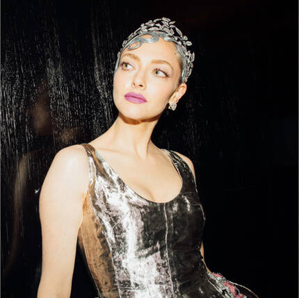 Una mujer con un vestido plateado brillante, adornado con flores, y una diadema de piedras brillantes, mira hacia un lado con una expresion elegante.