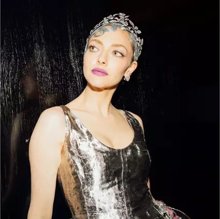 Una mujer con un vestido plateado brillante, adornado con flores, y una diadema de piedras brillantes, mira hacia un lado con una expresion elegante.