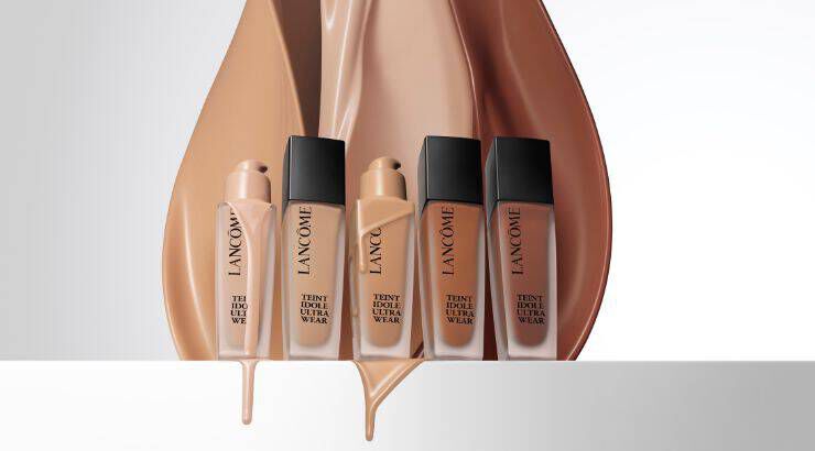 Imagen de varios frascos de base de maquillaje de la marca Lancome, mostrando diferentes tonalidades junto a fluido de maquillaje derramado en colores nude.