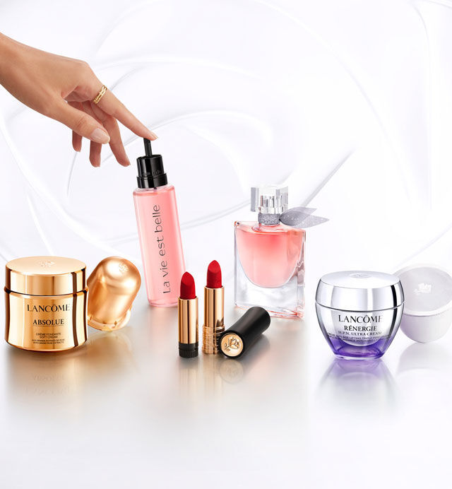 Imagen que muestra varios productos de belleza de la marca Lancome, incluyendo fragancias, labiales y cremas, dispuestos sobre una superficie brillante. Se observa una mano sosteniendo un frasco de suero.