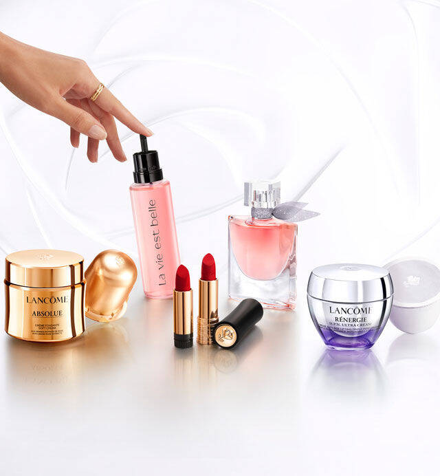 Imagen que muestra varios productos de belleza de la marca Lancome, incluyendo fragancias, labiales y cremas, dispuestos sobre una superficie brillante. Se observa una mano sosteniendo un frasco de suero.