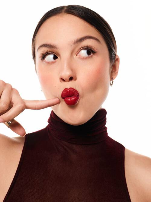 Mujer con maquillaje brillante y labios rojos, haciendo una expresion divertida mientras senala su boca con un dedo.