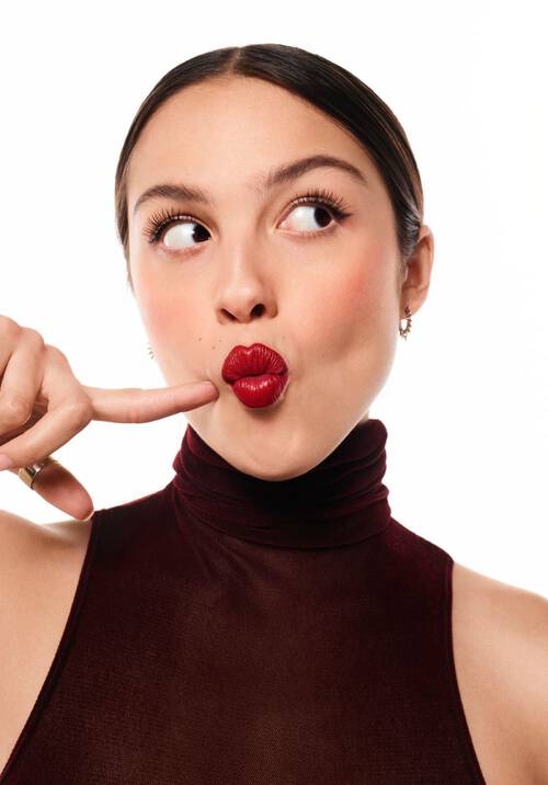 Joven con labios rojos brillantes, haciendo un gesto de "shhh" con un dedo en los labios, vestida con un top de cuello alto oscuro y una expresion juguetona.