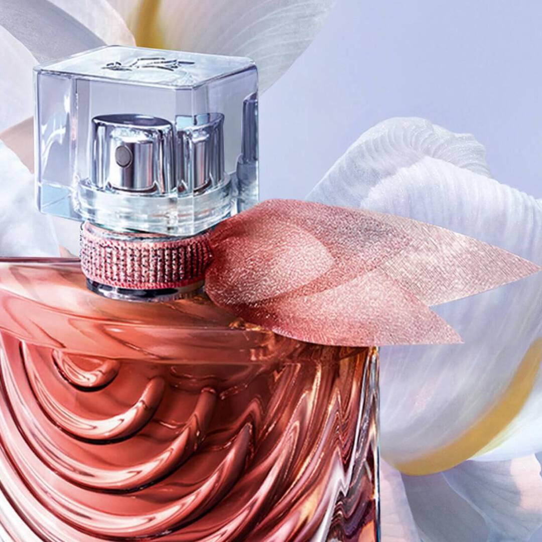 Botella de perfume elegante con un diseno suave en tonos rosados, adornada con un lazo de tela brillante y un fondo de flores.