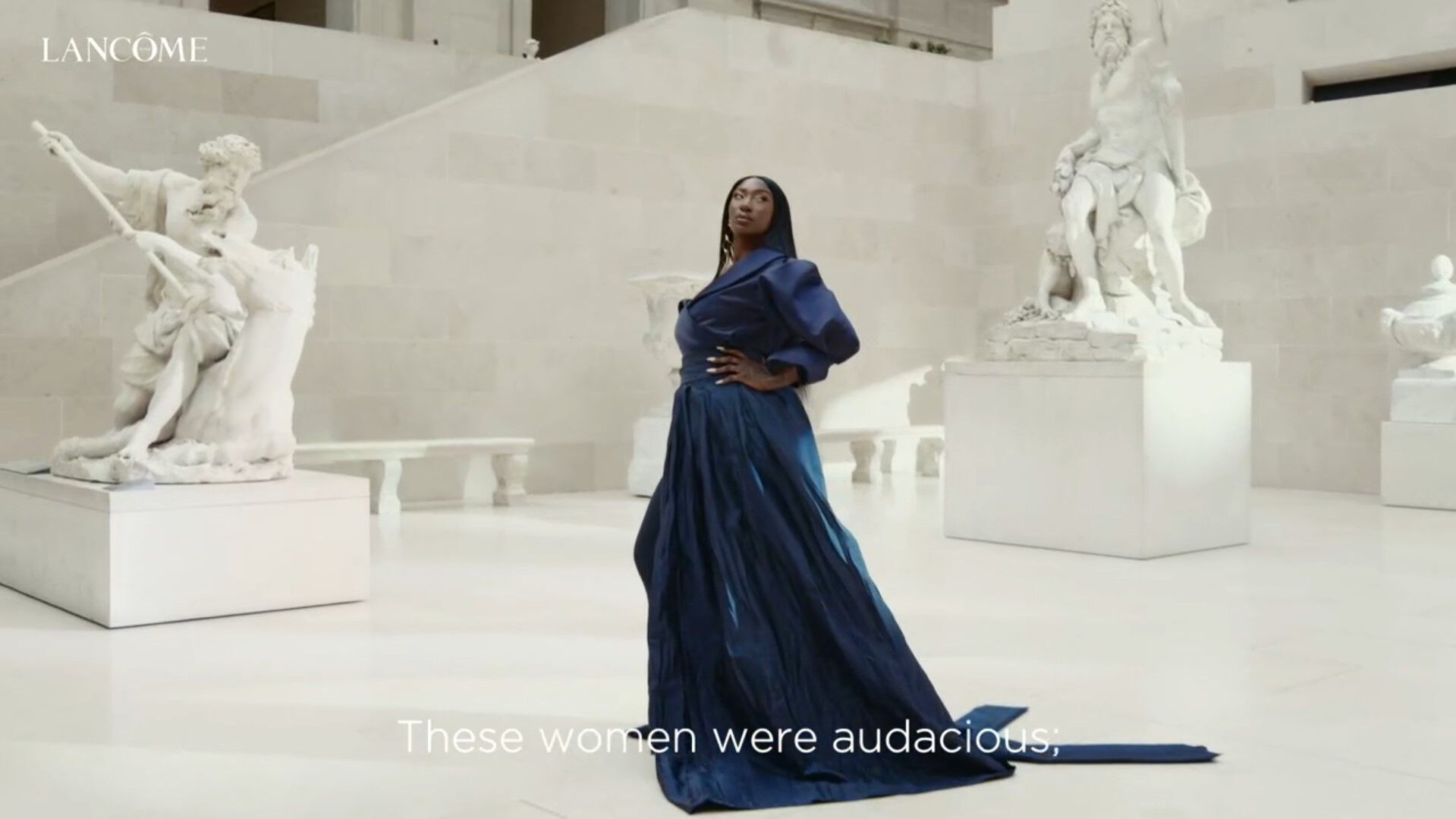 Una mujer de pie en un museo, vestida con un elegante vestido azul, rodeada de esculturas clasicas.