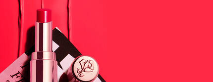 Un labial rosa brillante sobre una superficie de color rojo, acompanado de un envase elegante con el logotipo de la marca.