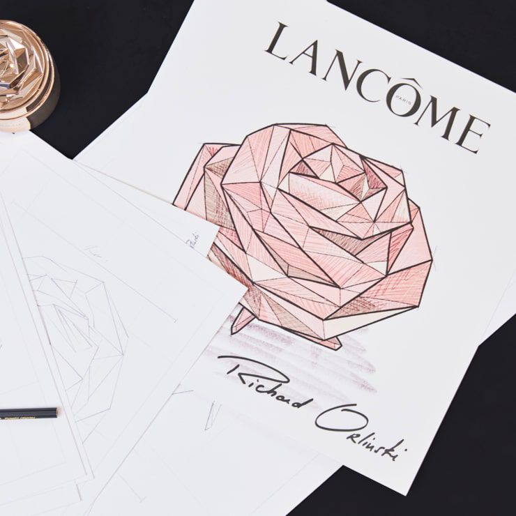 Ilustracion de una rosa geometrica en tonos rosados con el logo de Lancome y la firma de Richard Orlinski, sobre un fondo de hojas de papel.