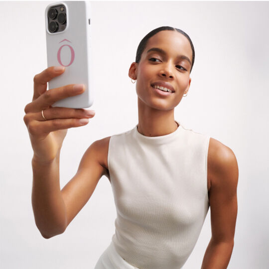 Una mujer sonriente toma una selfie con su telefono movil, luciendo una camiseta de manga corta en color claro.
