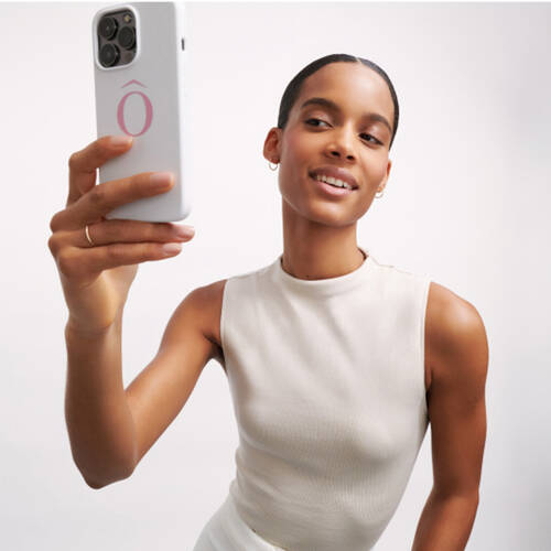 Una mujer sonriente toma una selfie con su telefono movil, luciendo una camiseta de manga corta en color claro.