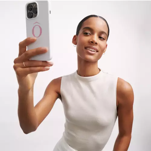Una mujer sonriente toma una selfie con su telefono movil, luciendo una camiseta de manga corta en color claro.