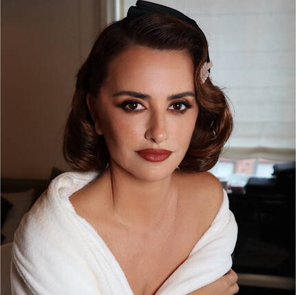 Mujer con cabello ondulado y maquillaje elegante, vestida con una bata blanca, posando con una expresion suave.