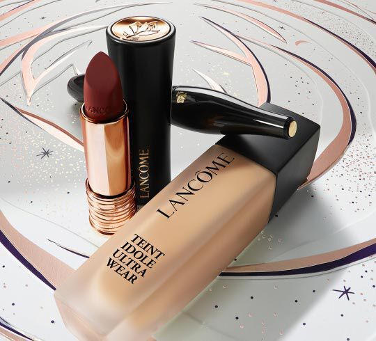 Imagen que muestra productos de maquillaje de Lancome, incluyendo un lapiz labial de color rojo oscuro y una base de maquillaje en un frasco elegante, sobre un fondo decorativo.