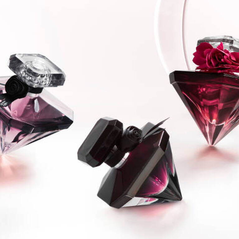 Frascos de perfumes de formas geometricas en tonos purpura y negro, con detalles de flores en la parte superior, sobre una superficie blanca.