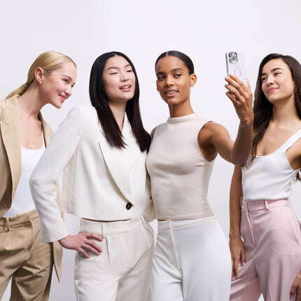 Cuatro mujeres de diferentes etnias posan juntas, sonriendo y tomando una selfie. Visten trajes modernos en tonos claros.
