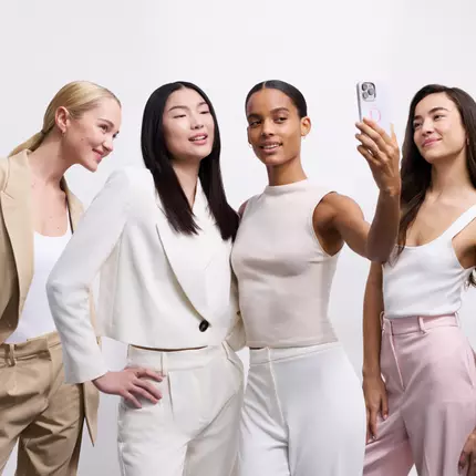 Cuatro mujeres de diferentes etnias posan juntas, sonriendo y tomando una selfie. Visten trajes modernos en tonos claros.