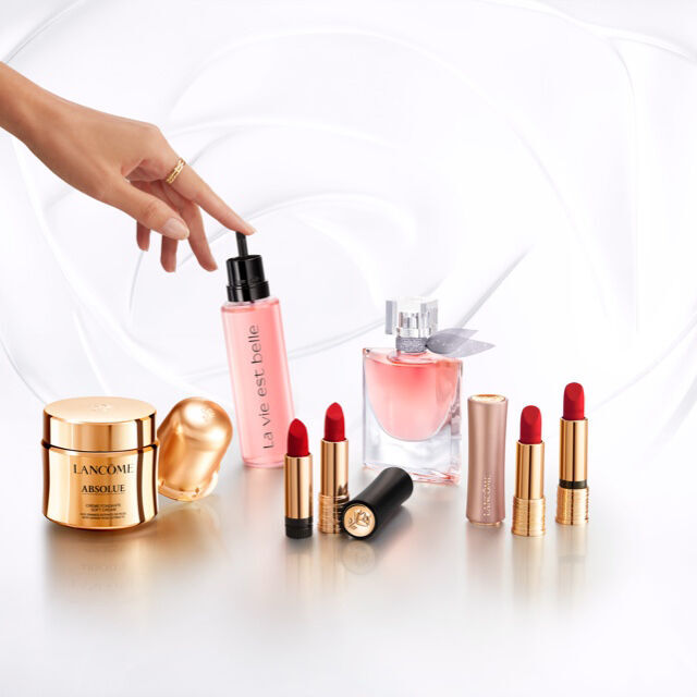 Imagen de productos de maquillaje y cuidado de la piel, incluidos labiales rojos, perfume y una crema hidratante, con una mano senalando un frasco rosa.