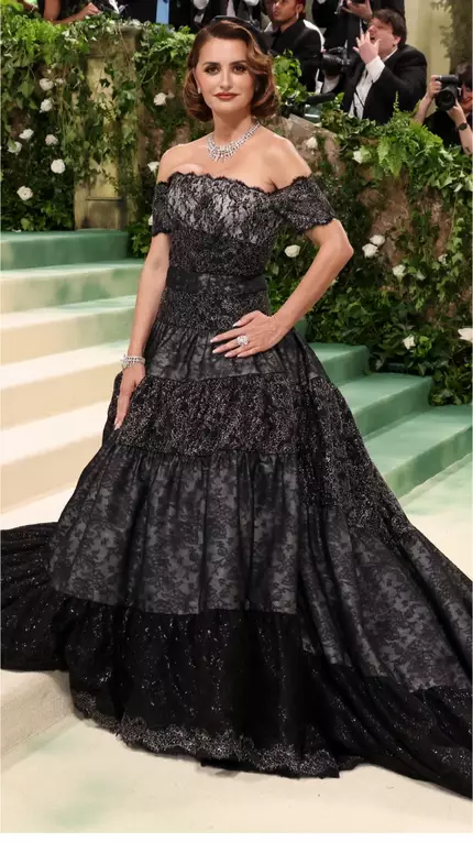 Una mujer en un elegante vestido negro con detalles brillantes, posando en una alfombra roja rodeada de fotografos y un ambiente floral.