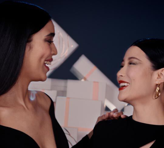 Dos mujeres sonriendo y mirandose con alegria, con un fondo de cajas decorativas.