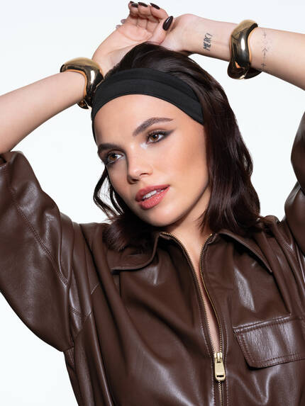 Mujer con cabello corto y oscuro, usando una camiseta de cuero marron y una cinta negra en la cabeza, posando con una expresion confiada.