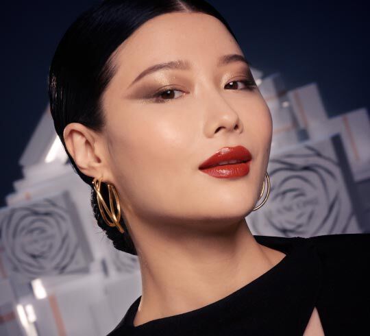 Retrato de una mujer con piel clara, labios rojos brillantes y maquillaje elegante, usando pendientes dorados y con el cabello recogido.