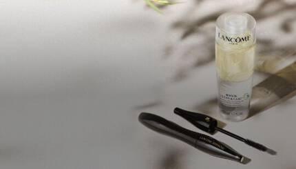 Botella de limpiador y cuidado de Lancome junto a un aplicador de mascara de pestanas sobre una superficie clara y decorada con hojas.