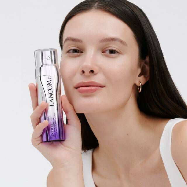 Mujer sosteniendo un frasco de producto de belleza, sonriente, con fondo blanco.